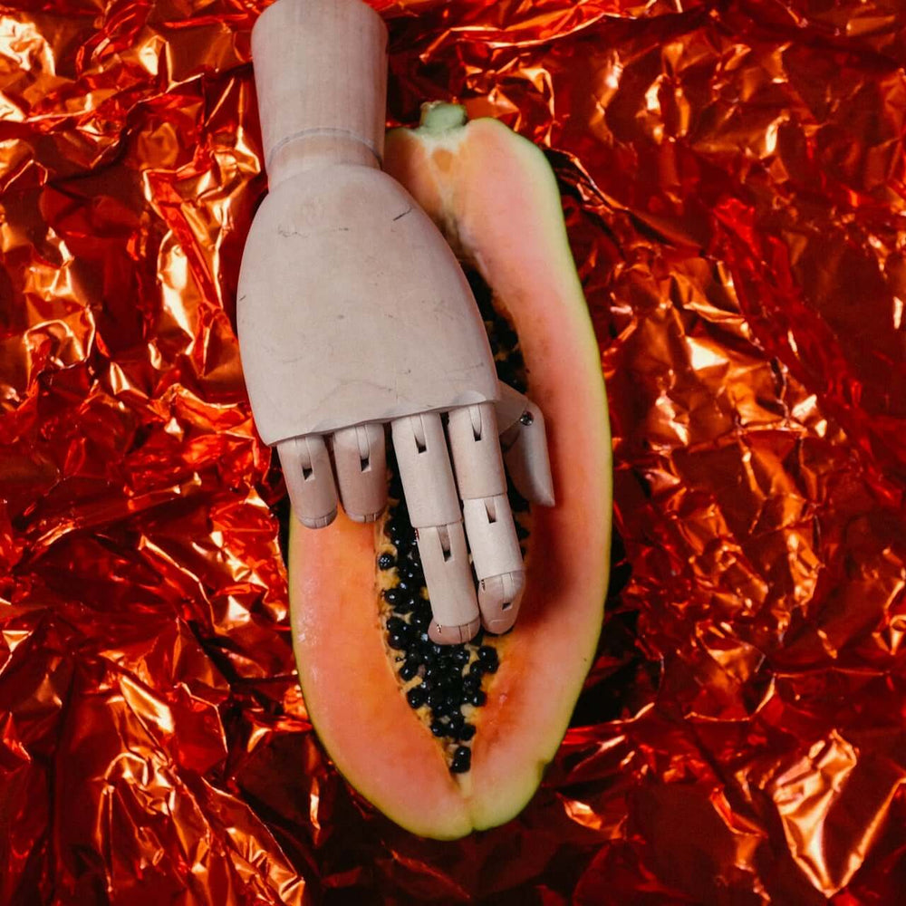 ALT-Text: "Eine hölzerne Mannequin-Hand liegt auf einer aufgeschnittenen Papaya auf einem roten metallischen Hintergrund, symbolisch für die intime Berührung im Kontext von CBD und Intimität."