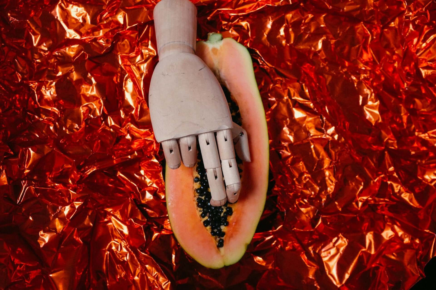 ALT-Text: "Eine hölzerne Mannequin-Hand liegt auf einer aufgeschnittenen Papaya auf einem roten metallischen Hintergrund, symbolisch für die intime Berührung im Kontext von CBD und Intimität."